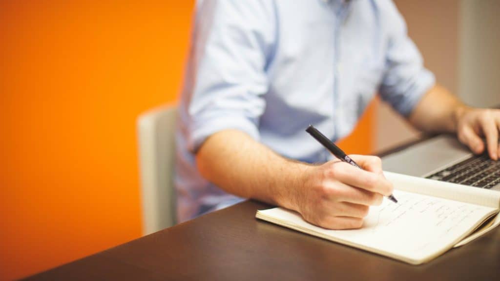 A photo of an individual working at a laptop while writing in a notebook.