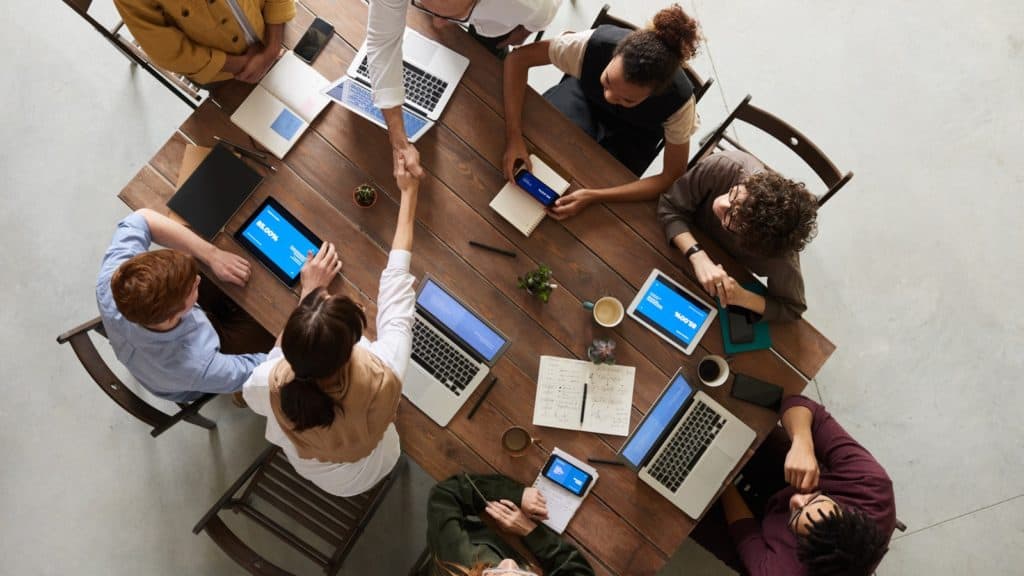 A photo looking down at a group of professionals, seated at a wood table, shaking hands and using their laptops and mobile devices.