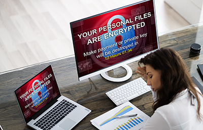 A photo a woman sitting at a desk in front of a laptop and monitor showing a ransomware message.