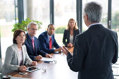A photo of a businessman speaking in front of a group of people at a conference table.